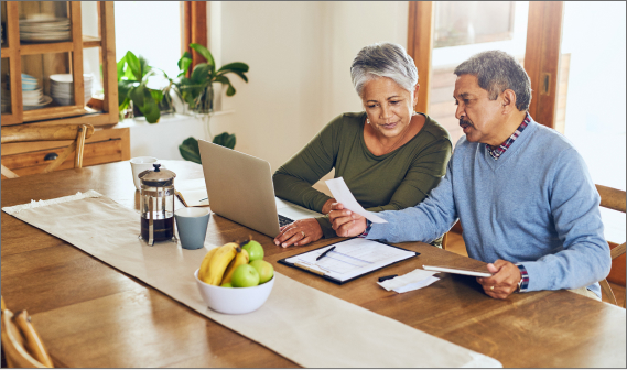 Two people sitting at a dining table. One person has a laptop and another is looking at a piece of paper.